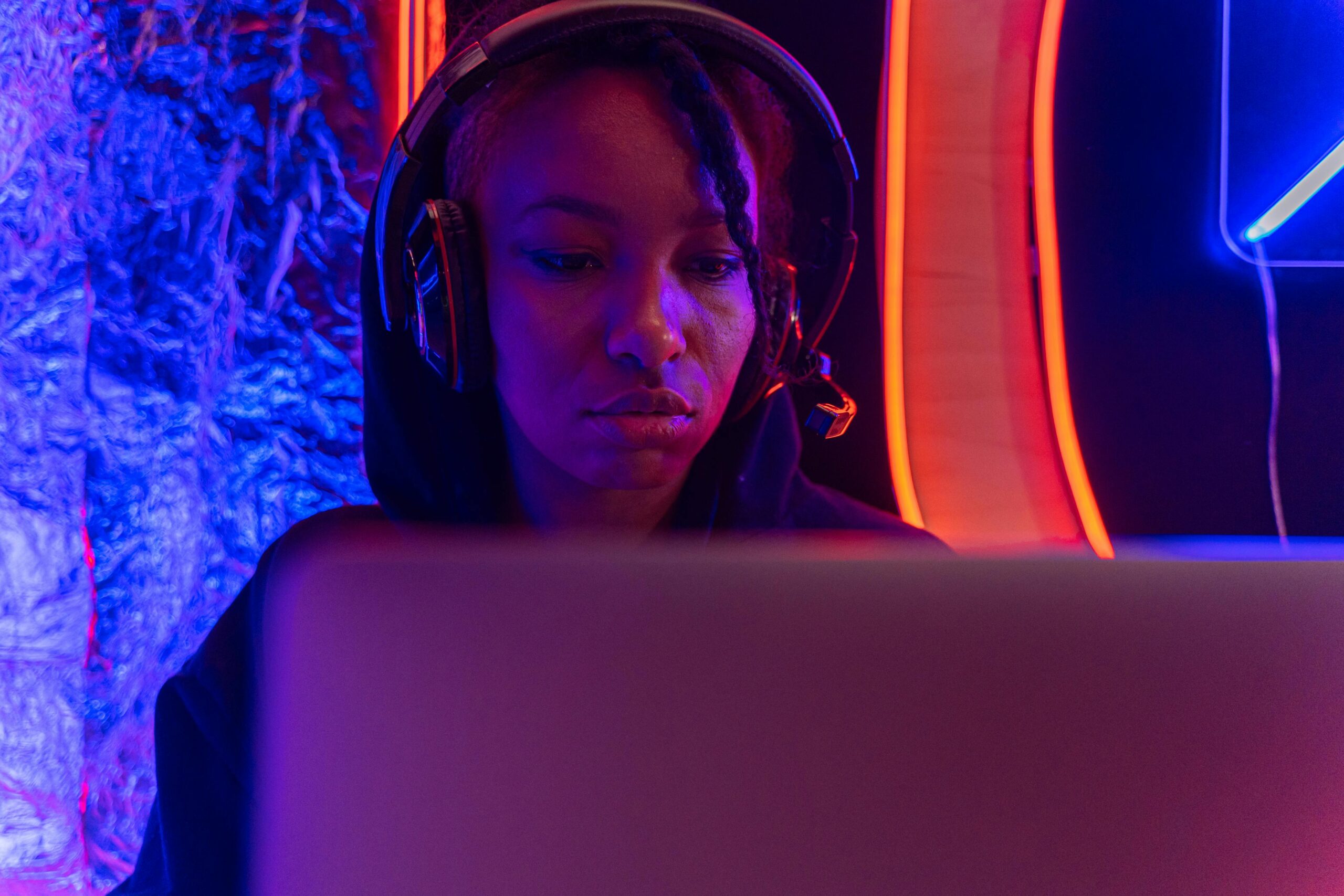 A woman wearing headphones focused on her laptop in a dimly lit room with neon lights.